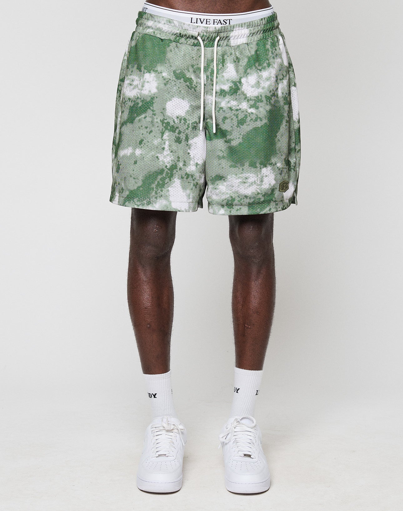 A person wears LFDY Mesh Shorts by LIVE FAST DIE YOUNG in green and white tie-dye with an elastic waistband featuring LIVE FAST, paired with white socks and sneakers, standing against a plain white background.