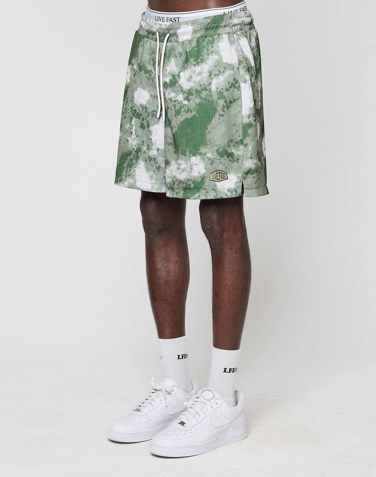 A person wears green and white LFDY Mesh Shorts by LIVE FAST DIE YOUNG (Regular Fit, elastic waistband), white LIVE FAST socks, and white Nike sneakers. Only their legs and lower torso are shown against a plain white background.