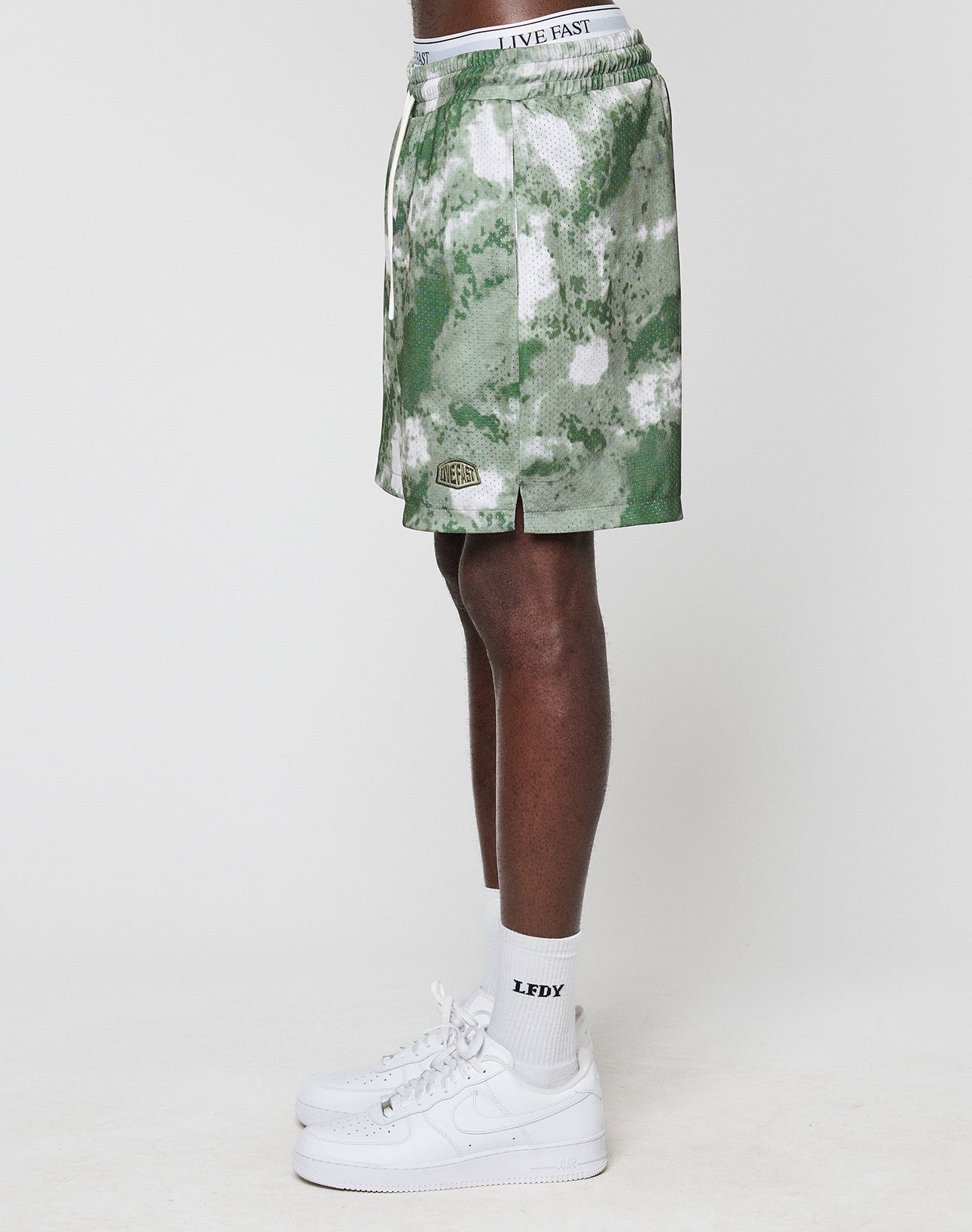A person stands sideways against a plain background wearing LIVE FAST DIE YOUNG LFDY Mesh Shorts with a green camo pattern, elastic waistband featuring LIVE FAST text, regular fit, plus white LFDY socks and Nike Air Force 1 sneakers.
