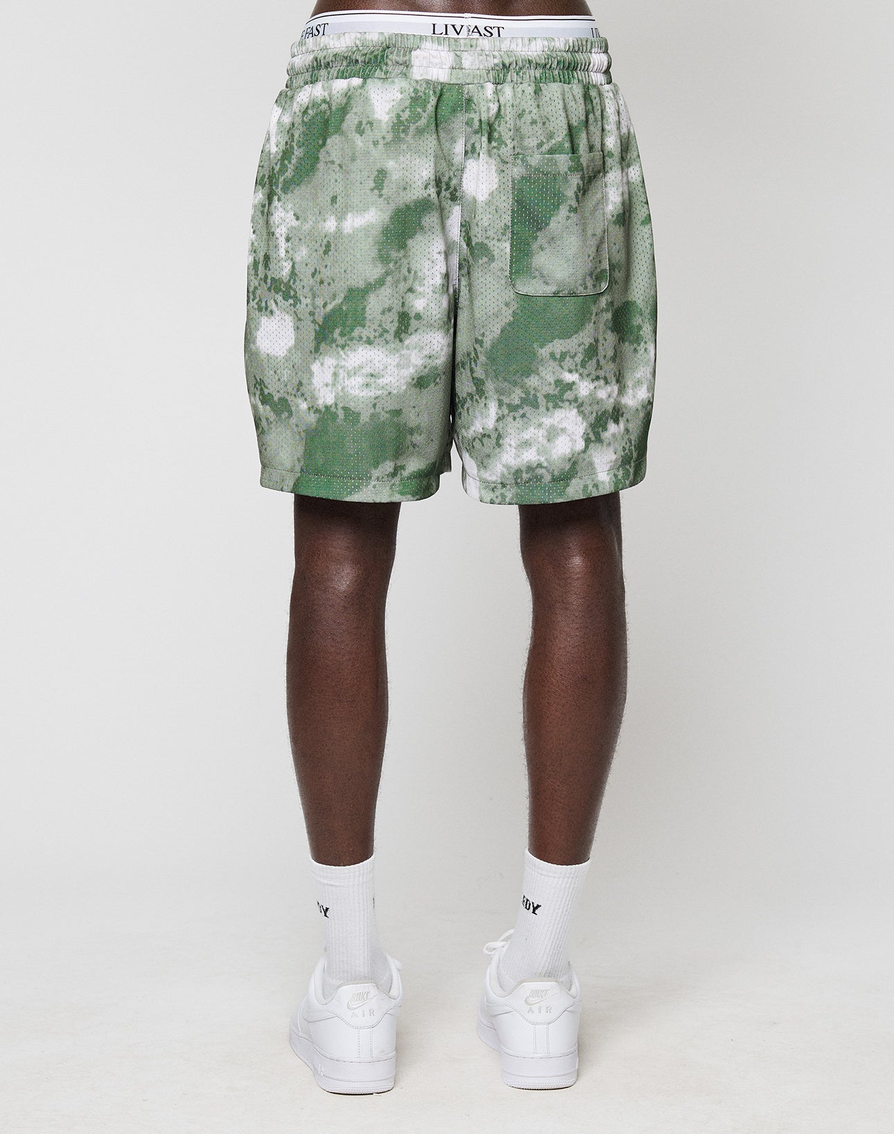 Someone stands with their back to the camera, wearing green and white LFDY Mesh Shorts by LIVE FAST DIE YOUNG, white socks, white sneakers, and a white waistband underwear, all set against a plain white background.