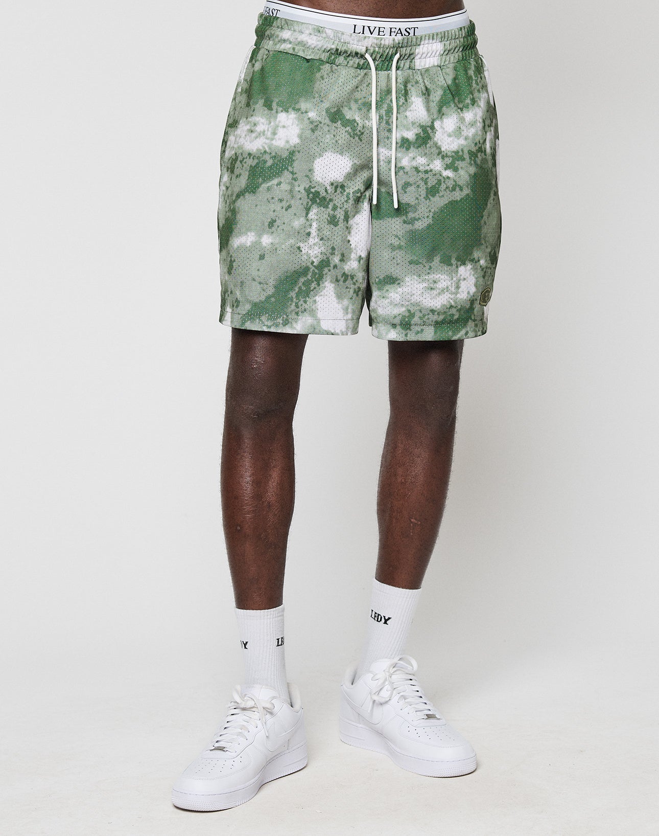 A person wearing green and white LFDY Mesh Shorts by LIVE FAST DIE YOUNG, featuring a LIVE FAST elastic waistband and drawstring, paired with white socks and sneakers, stands against a plain white background. The shorts are soft polyester.