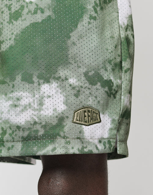 Close-up of a person wearing LFDY Mesh Shorts by LIVE FAST DIE YOUNG. The green and white camo polyester shorts have an elastic waistband, regular fit, and a small LIVE FAST patch at the hem, revealing part of their dark-skinned leg.