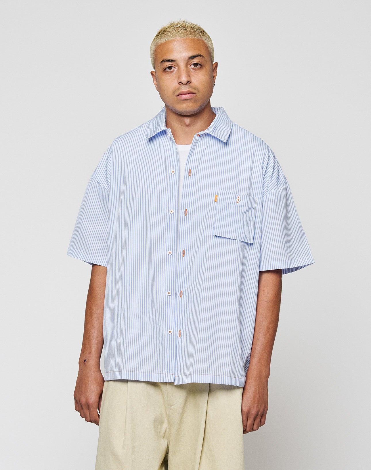 A person with short blond hair wears the LIVE FAST DIE YOUNG LFDY Striped Boxy Shirt in light blue and white, featuring a subtle logo stitch, paired with beige pants. They stand against a plain light gray background.
