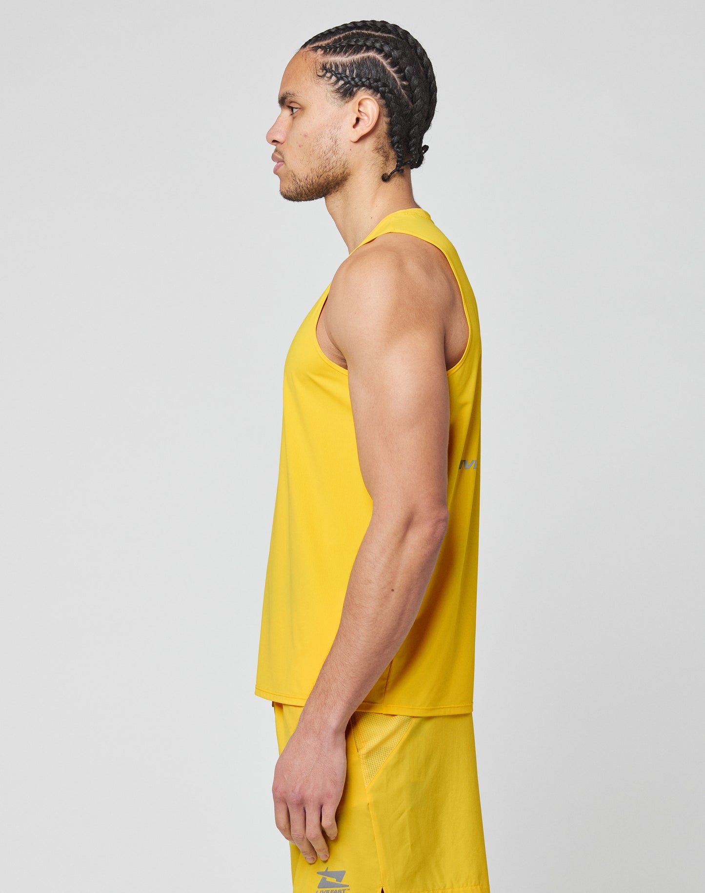 A man with braided hair stands in profile, wearing a slim fit, sleeveless yellow Live Fast Performance Tank and matching yellow shorts by LIVE FAST DIE YOUNG against a plain light gray background.