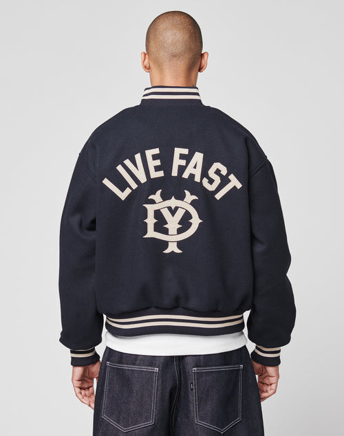 A person with a shaved head wears the LIVE FAST DIE YOUNG Live Fast College Jacket featuring beige trim, a DY-Reißverschluss Puller, large beige LIVE FAST text, a stylized LY logo on the back, and a classic regular fit.