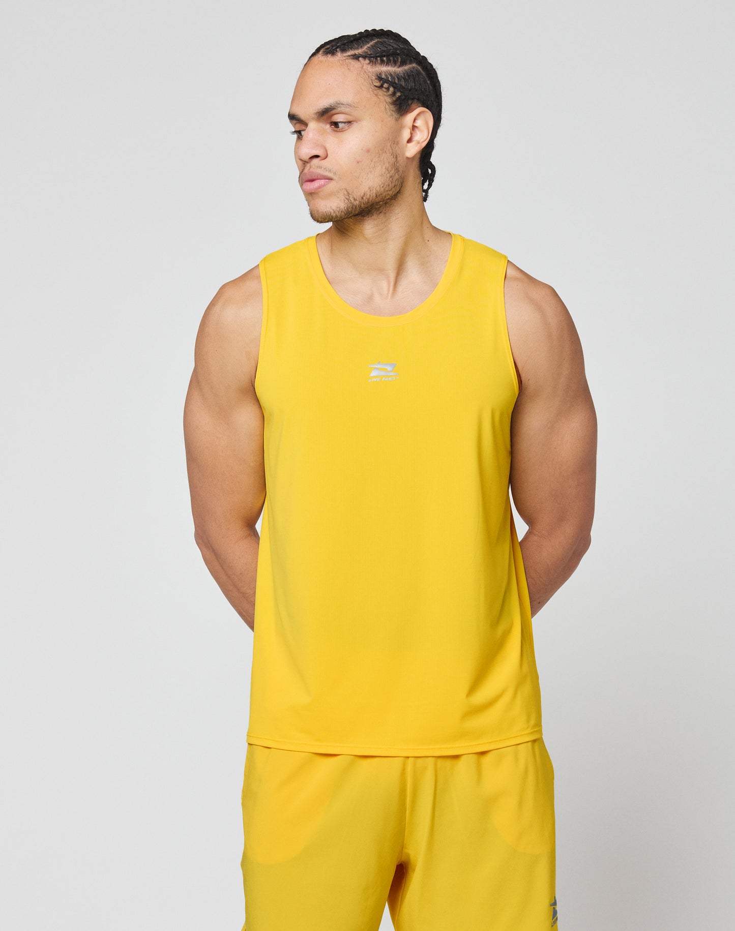 A man models the LIVE FAST DIE YOUNG Live Fast Performance Tank in bright yellow with matching shorts featuring reflective print, standing against a plain light gray background and looking left with his hands behind his back.