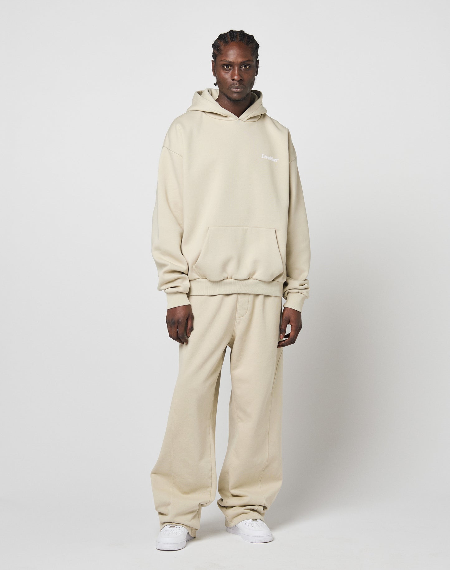 A person in a loose beige Basic Wide Jogger by LIVE FAST DIE YOUNG with an elastic waistband stands against a white background. They have short braided hair and wear white sneakers.