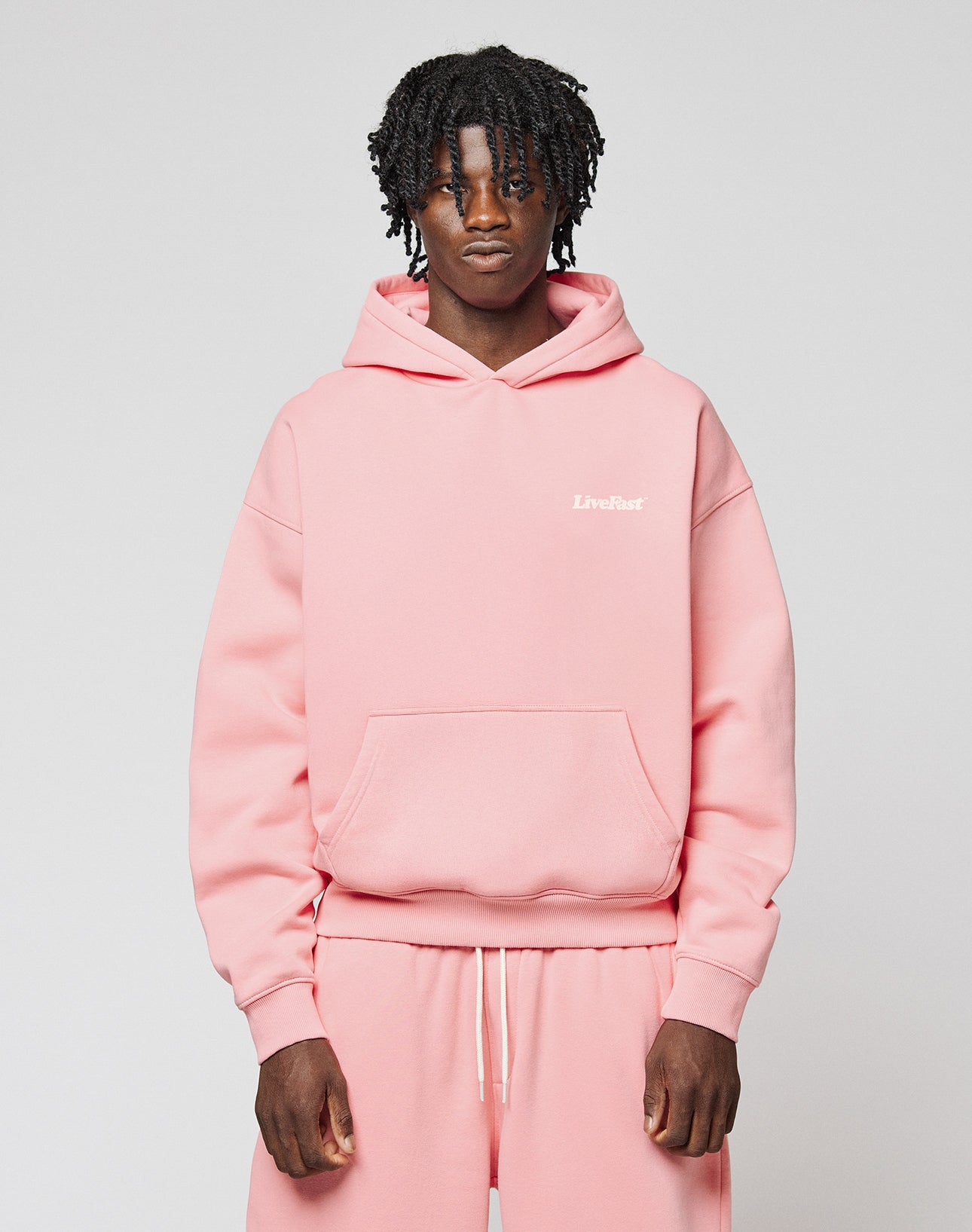 A person with short, curly hair wears an oversize fit light pink Basic 360 Hooded sweatshirt and matching sweatpants from LIVE FAST DIE YOUNG, featuring subtle topstitching and “LiveFast” in small white letters on the chest. The background is plain.