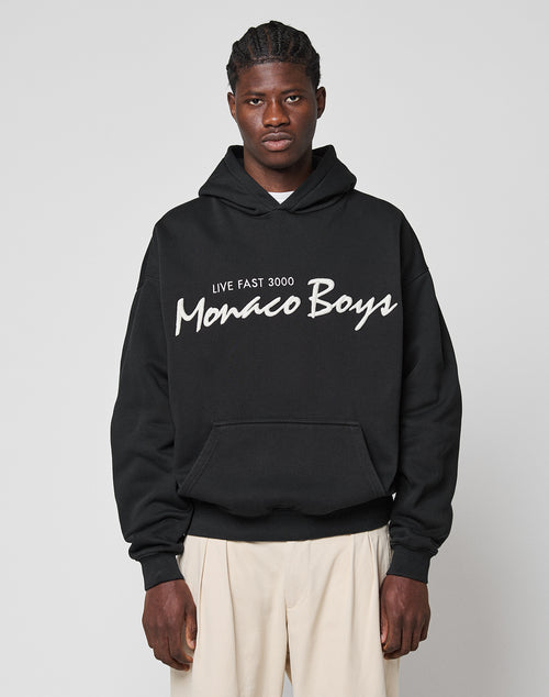 A man wears the LIVE FAST DIE YOUNG Monaco Boys Hooded in an oversize fit with a 3D LIVE FAST 3000 Monaco Boys graphic on the front, paired with beige cotton-polyester mix pants, standing against a plain light gray background.