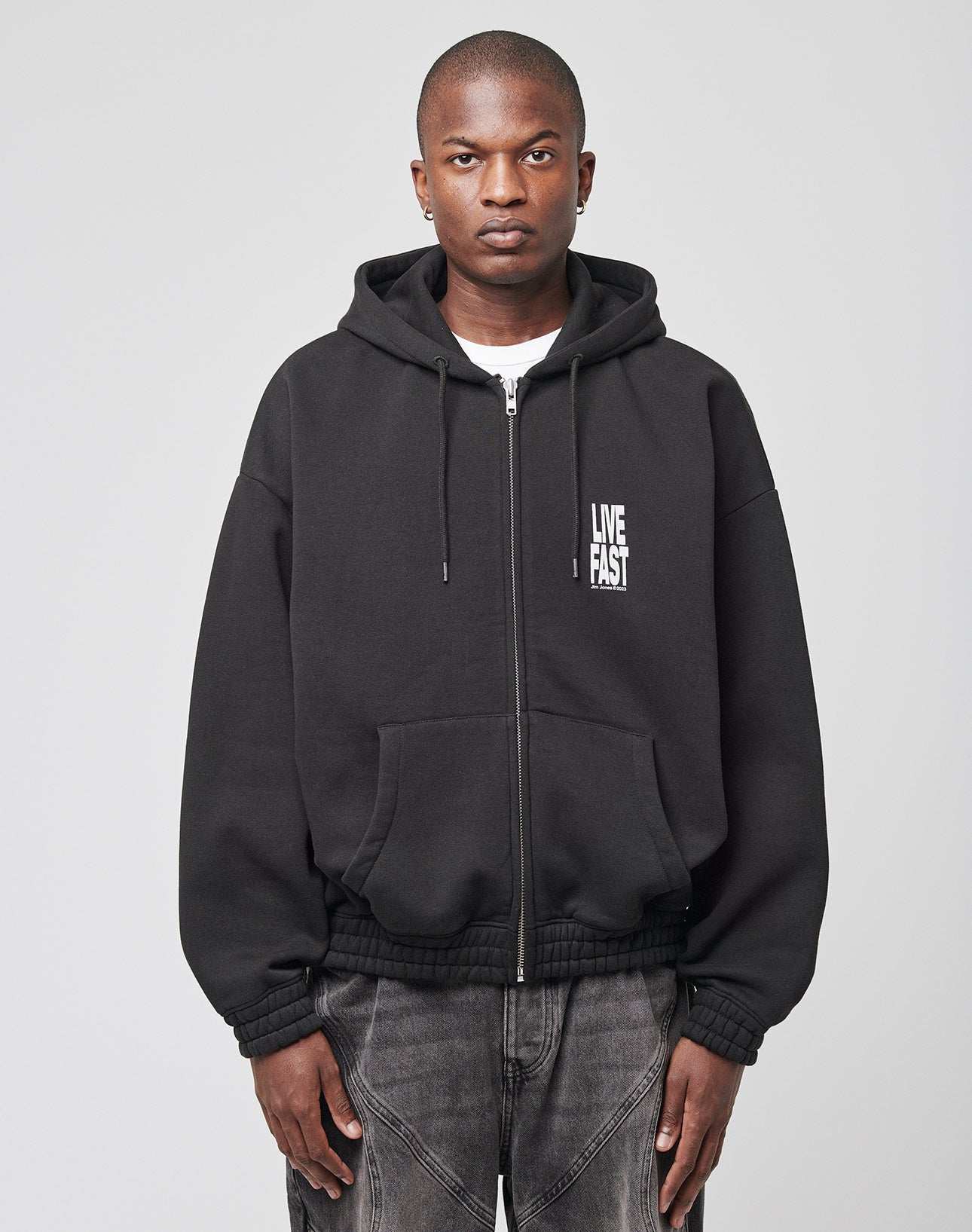 Against a plain light gray background, a person wears the LIVE FAST DIE YOUNG Live Fast Lenox Hooded Zip—an oversize fit black zip-up hoodie with LIVE FAST in white on the chest—paired with faded black jeans. Hands at sides, they have a neutral expression.