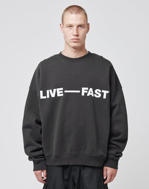A person with short blond hair wears the LIVE FAST DIE YOUNG Live – Fast Sweater, an oversize fit black sweatshirt with LIVE — FAST in bold white letters, standing against a plain light gray background.