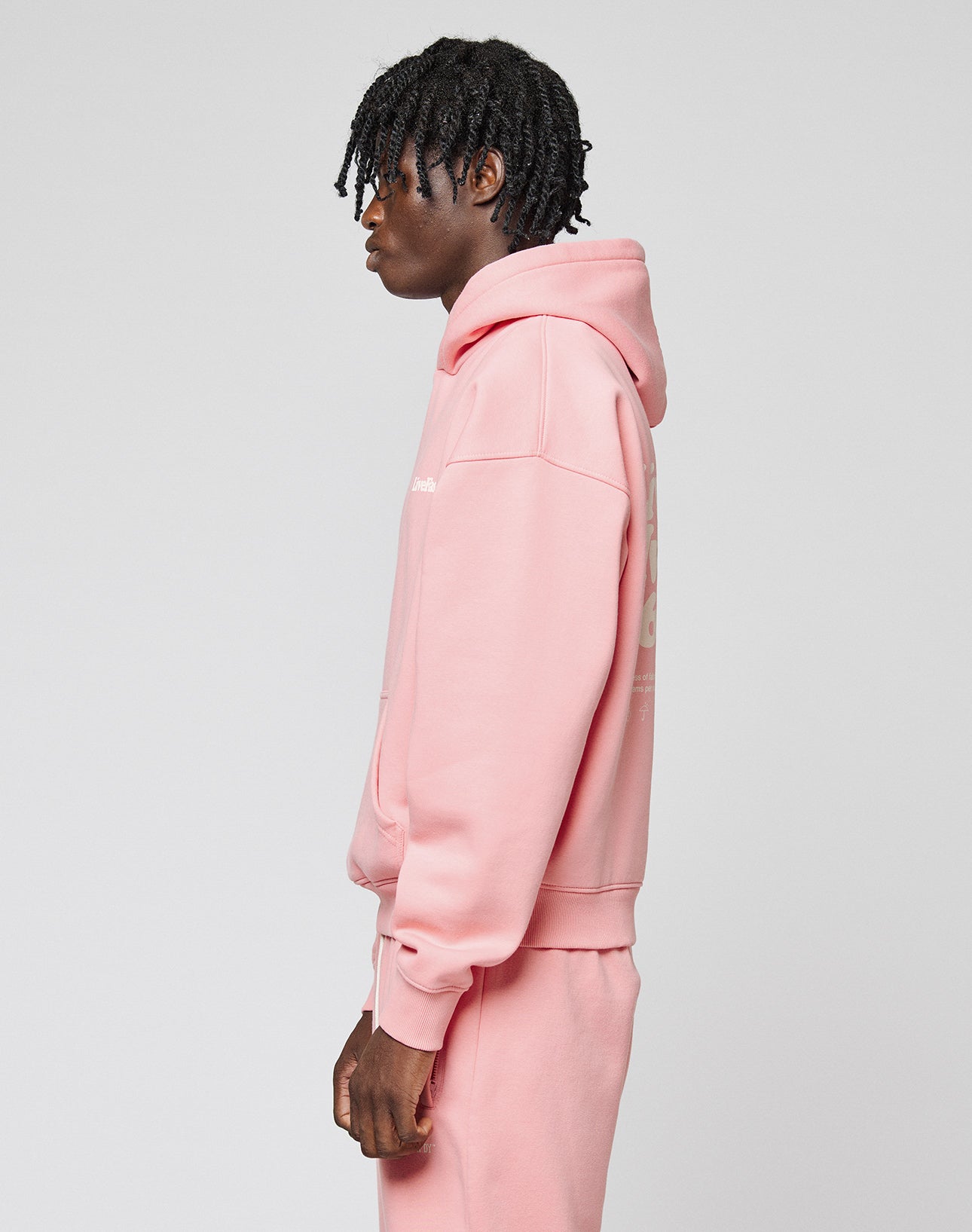A person with short, twisted hair wears the oversize fit Basic 360 Hooded by LIVE FAST DIE YOUNG in pink with matching sweatpants, featuring topstitching, standing in profile against a plain light gray background.