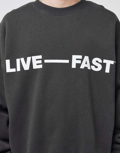 A person wears an Oversize Fit Live – Fast Sweater by LIVE FAST DIE YOUNG, featuring bold white LIVE—FAST text across the chest. The image is cropped to the upper torso and part of the neck. Made from a comfortable Baumwolle blend.