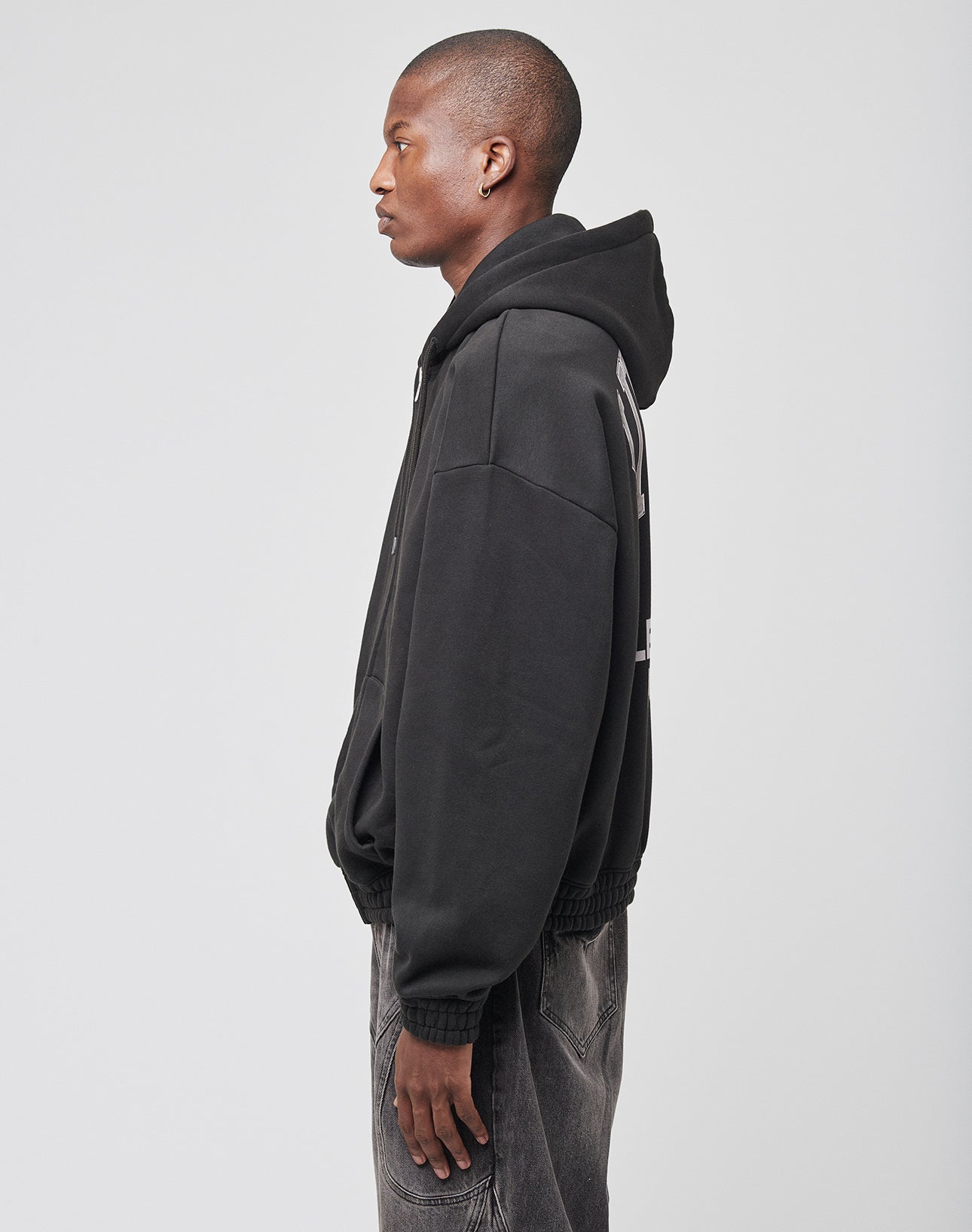 A person stands in profile facing left, wearing the LIVE FAST DIE YOUNG Live Fast Lenox Hooded Zip and faded black jeans against a plain white background.