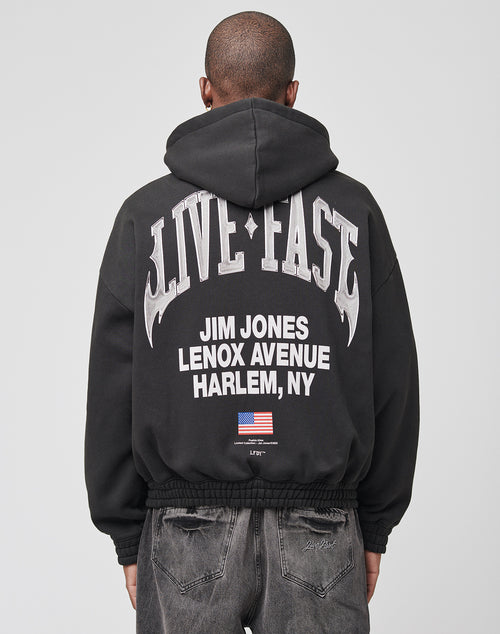 A person faces away from the camera wearing a black, oversized LIVE FAST DIE YOUNG Live Fast Lenox Hooded Zip, made of Baumwolle, with bold front and back prints including JIM JONES LENOX AVENUE HARLEM, NY and a small American flag on the back.