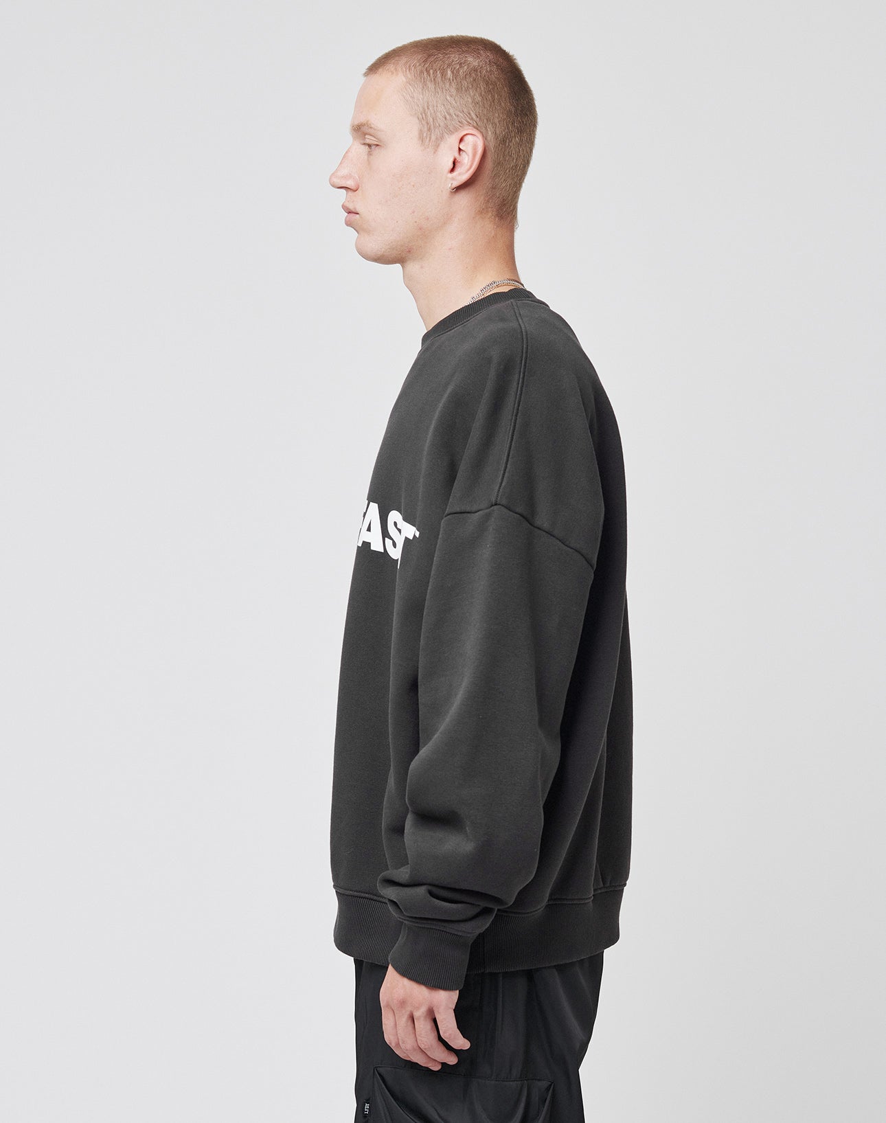 A person with short hair stands in profile against a plain background, wearing the black Live – Fast Sweater by LIVE FAST DIE YOUNG in an oversize fit with white text, made from Baumwolle and polyester, paired with black pants.