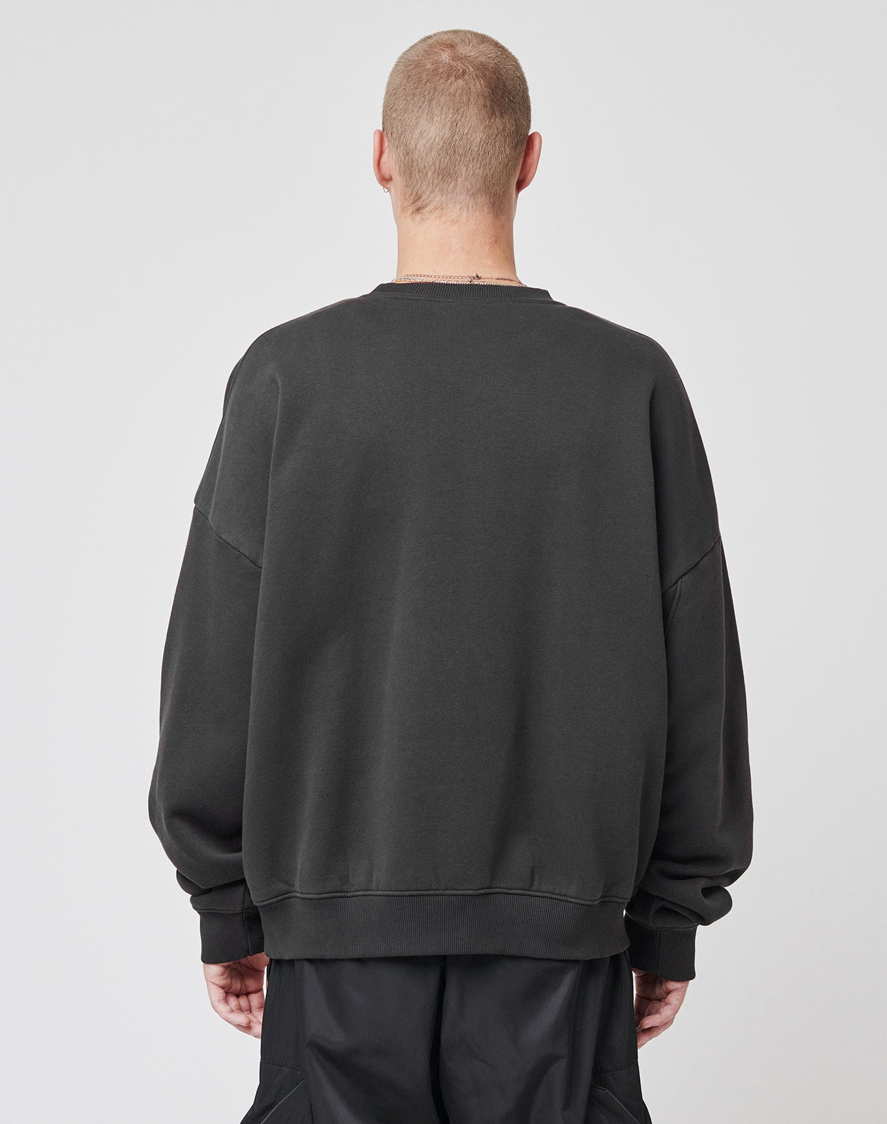 A person with short hair stands with their back to the camera, wearing the LIVE FAST DIE YOUNG Live – Fast Sweater, an oversized dark soft Baumwolle sweatshirt, paired with black pants against a plain light background.