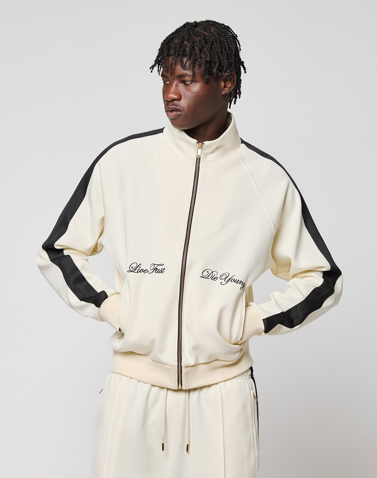 Someone stands against a plain background wearing the cream-colored LFDY Striped Track Jacket by LIVE FAST DIE YOUNG, featuring raglan sleeves, black stripes on the arms, and "Live Fast and Die Young" embroidered on the front.