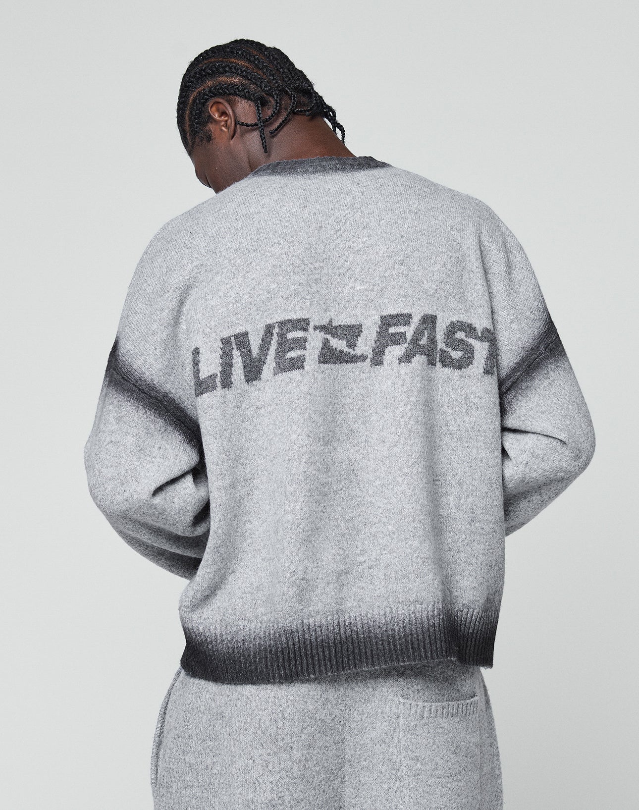 A person with braided hair faces away, wearing the LIVE FAST DIE YOUNG Live Fast Boucle Sweaterāan oversize gray piece featuring bold āLIVE FASTā lettering on the back. The background is plain and light-colored.