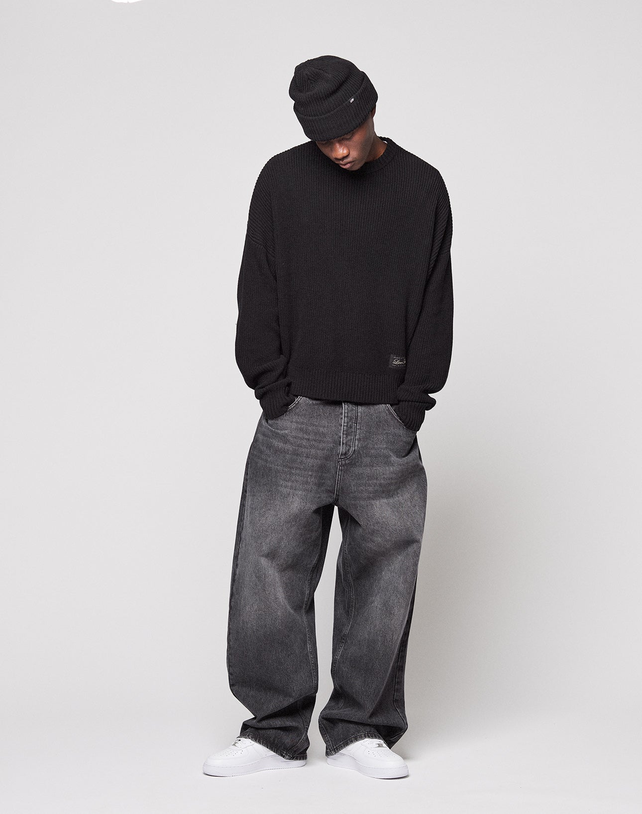 A person wears the LIVE FAST DIE YOUNG LFDY Knit Sweater with dropped shoulders and an oversize fit, paired with a black beanie, loose gray jeans, and white sneakers, standing against a plain light background looking down.