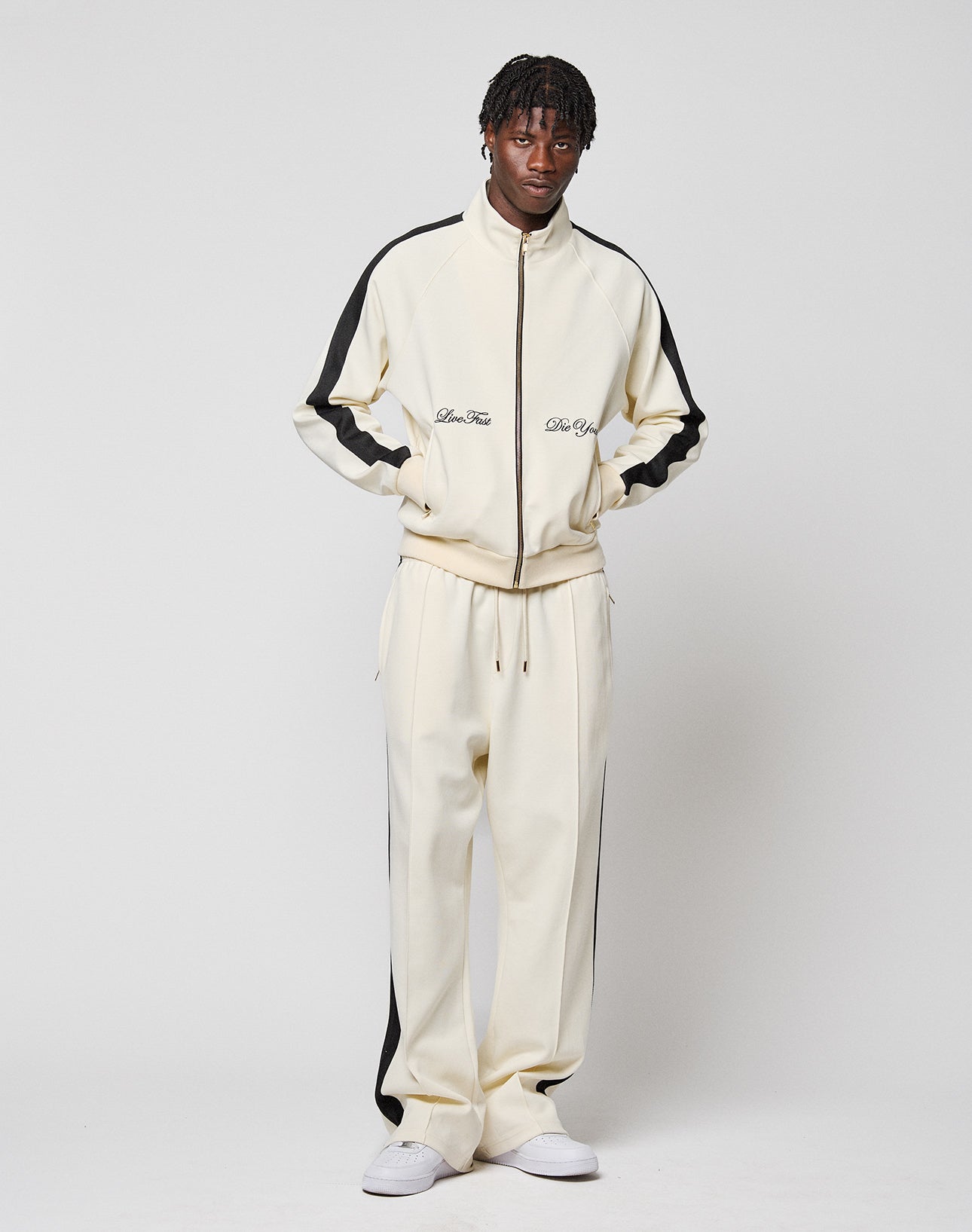 A man stands against a white background wearing the LIVE FAST DIE YOUNG LFDY Striped Jogger, a cream-colored tracksuit with black stripes and elastic waistband, paired with white sneakers and his hands in his pockets.