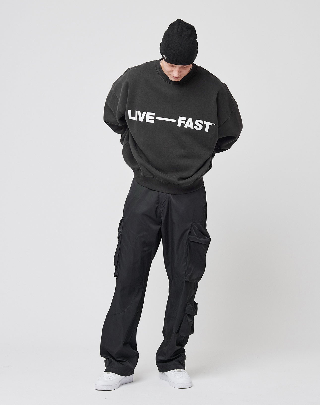 A person wearing a black beanie, black cargo pants, white sneakers, and the LIVE FAST DIE YOUNG Live – Fast Sweater featuring bold white “LIVE — FAST” lettering (Baumwolle/Polyester blend) stands against a plain white background, looking down.