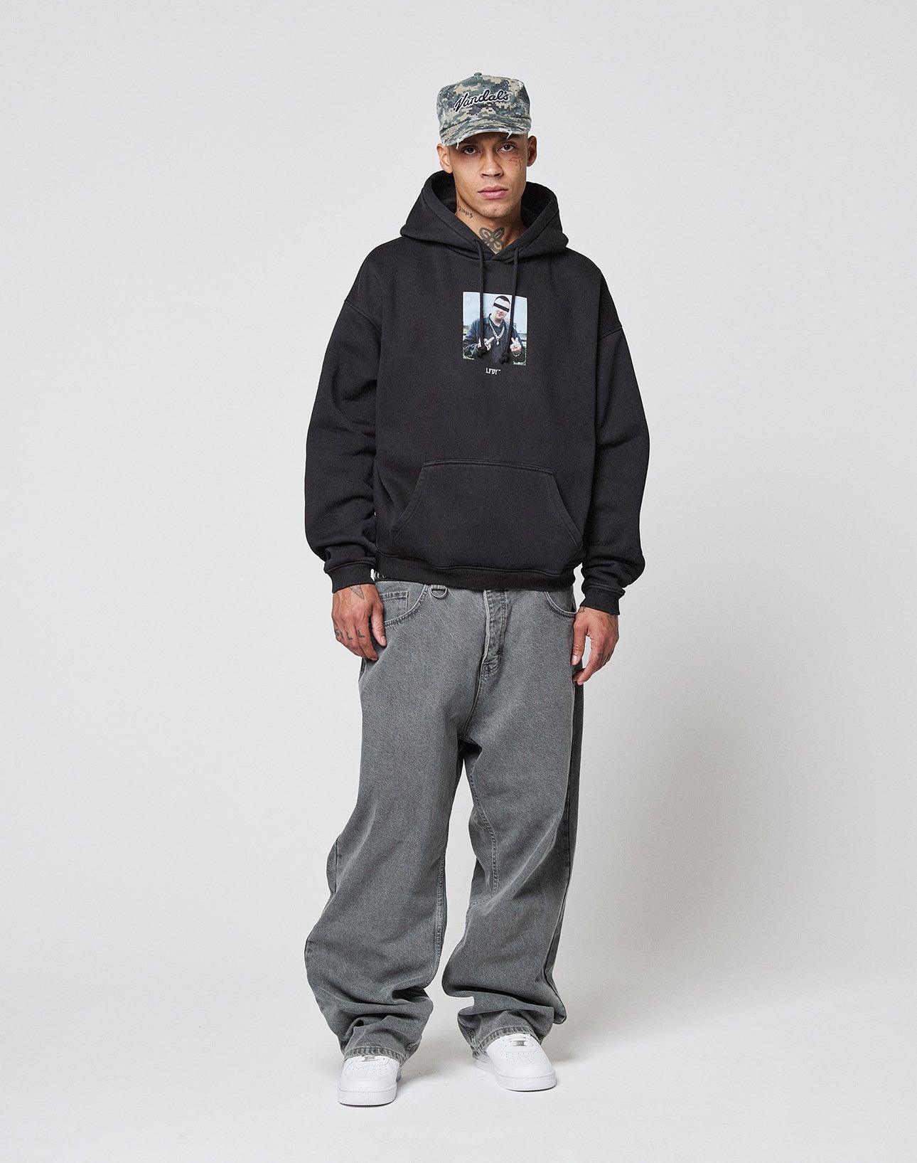 A man wearing a camo cap, LIVE FAST DIE YOUNG LFDY Gambling Hooded with frontprint, baggy gray topstitched jeans, and white sneakers stands against a plain white background.