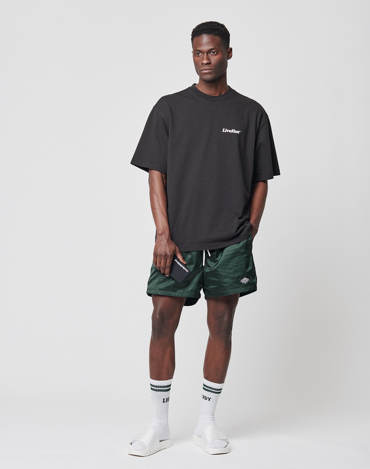 A man stands against a plain background wearing a black oversized t-shirt, LIVE FAST DIE YOUNG Basic Satin Shorts in green camo with Elastikbund, white socks with black stripes, white slide sandals, and holds a phone while looking to the side.