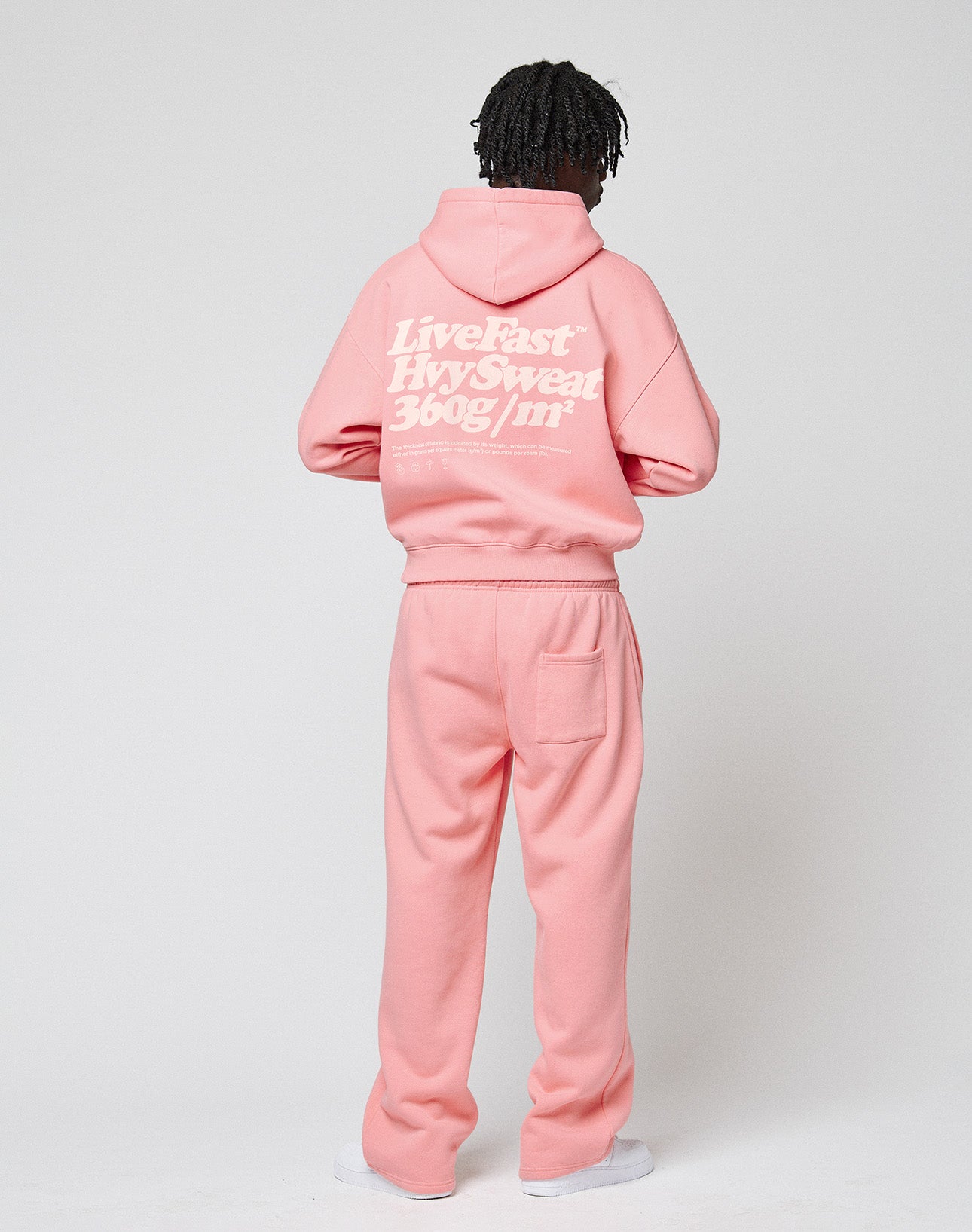 A person with short, twisted hair faces away, wearing an oversize light pink LIVE FAST DIE YOUNG Basic 360 Hooded sweatshirt and matching sweatpants. The hoodie features white “Live Fast– Hvysweat 360g/m²” text on the back. Plain white background.