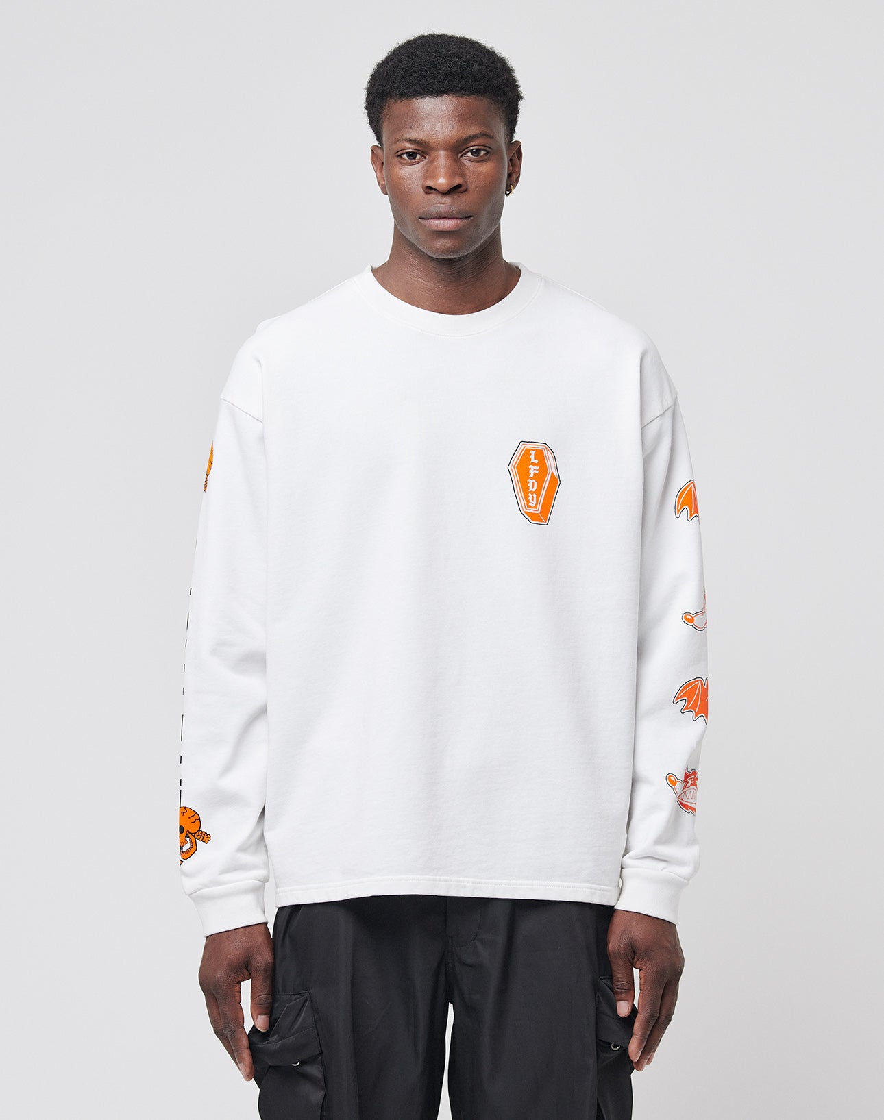 A man wearing the LIVE FAST DIE YOUNG No Rich Parents Longsleeveāa white regular fit shirt with orange graphics on the sleeves and chest, made from 100% Baumwolleāstands facing forward against a plain light gray background, paired with black pants.