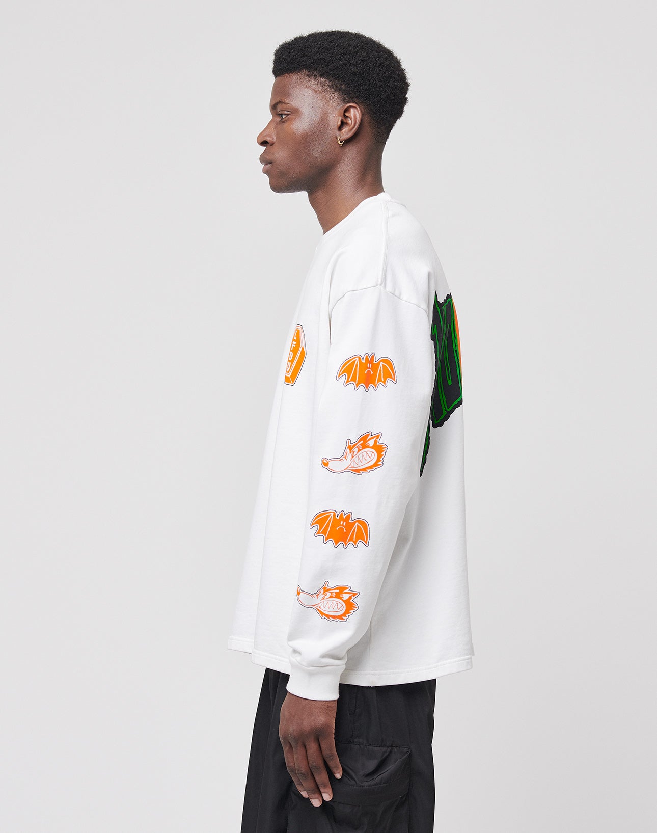 A young man stands in profile wearing the LIVE FAST DIE YOUNG No Rich Parents Longsleeve, a regular fit white 100% Baumwolle shirt with colorful dragon and logo patches on the sleeve, paired with black pants against a gray background.