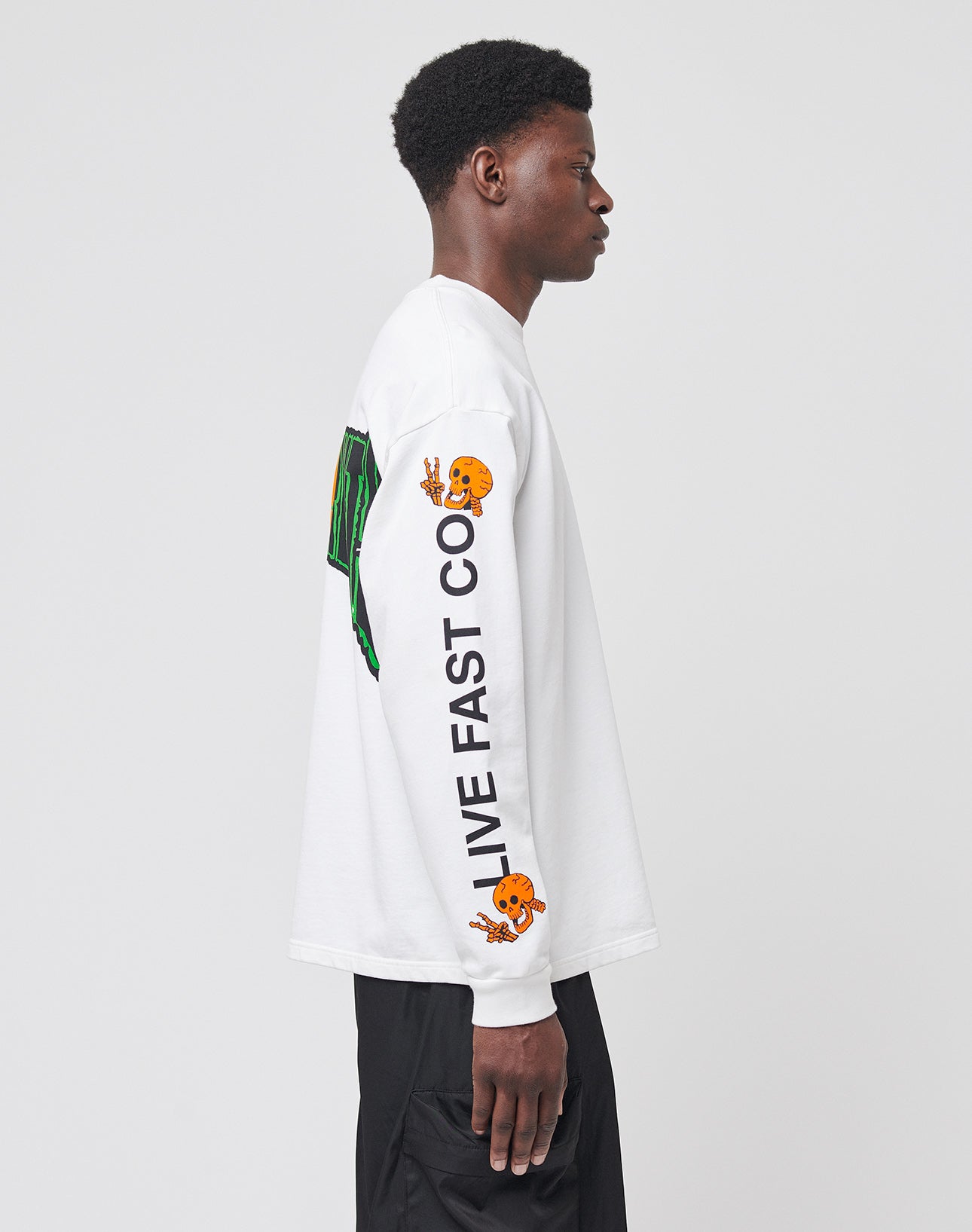 A person in profile wears the No Rich Parents Longsleeve by LIVE FAST DIE YOUNG, a white 100% Baumwolle shirt with black text LIVE FAST CO. and two orange cartoon graphics on the sleeve, paired with black pants against a light background.