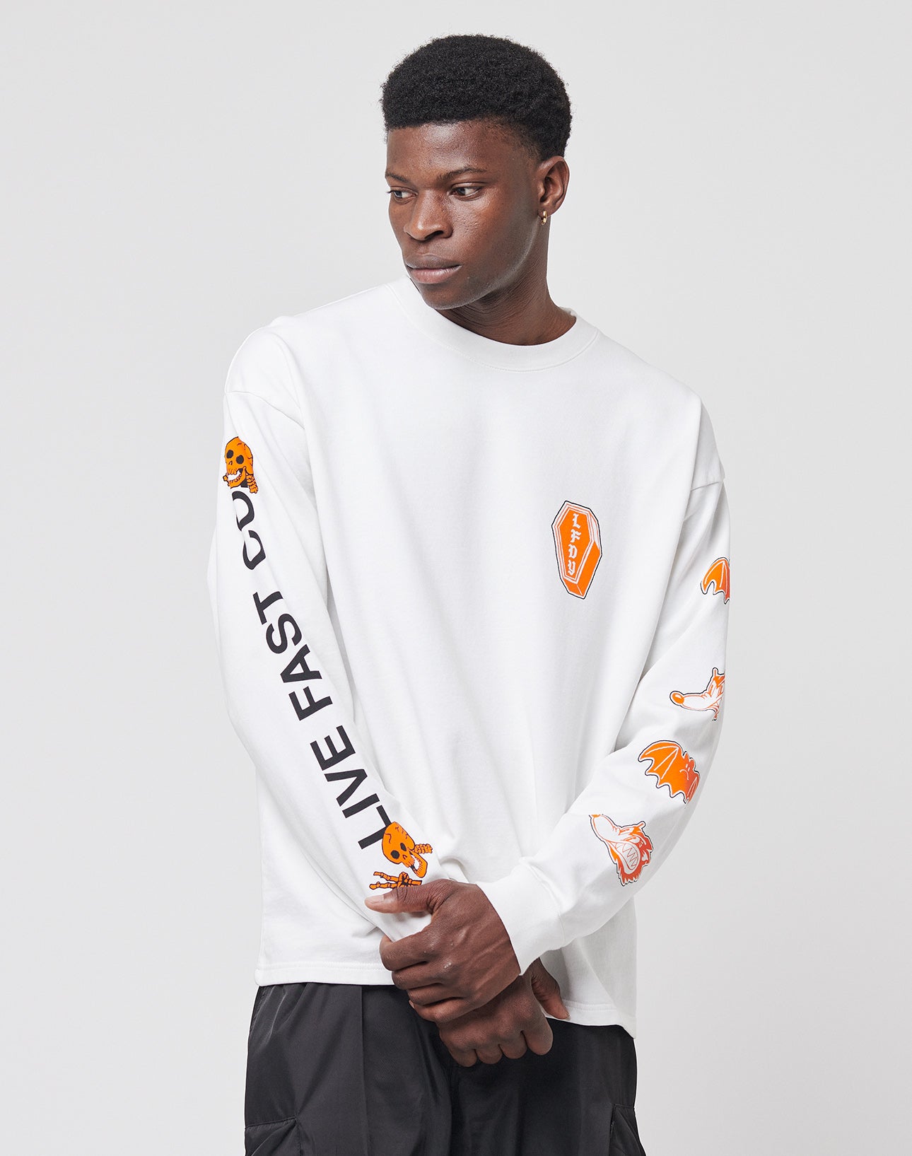 Someone wears the No Rich Parents Longsleeve by LIVE FAST DIE YOUNGāa regular fit, white 100% Baumwolle shirt with orange graphic patches and LIVE FAST in black on the left sleeveāarms crossed against a plain light background.