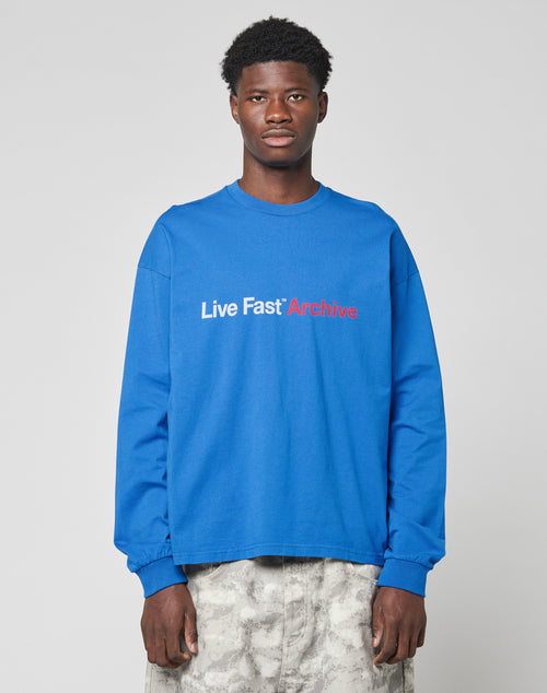 A person wears the LIVE FAST DIE YOUNG Live Fast Archive Longsleeve in bright blue with a bold white and red front print, made from 100% Baumwolle, styled with light gray patterned pants against a plain light gray background.