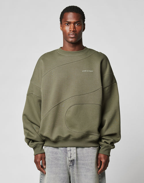 A man in the LFDY Wavy Sweater by LIVE FAST DIE YOUNG, an oversized olive green cotton-poly blend with subtle curved seams and light chest text, pairs it with wide-leg light jeans while standing against a plain gray background.
