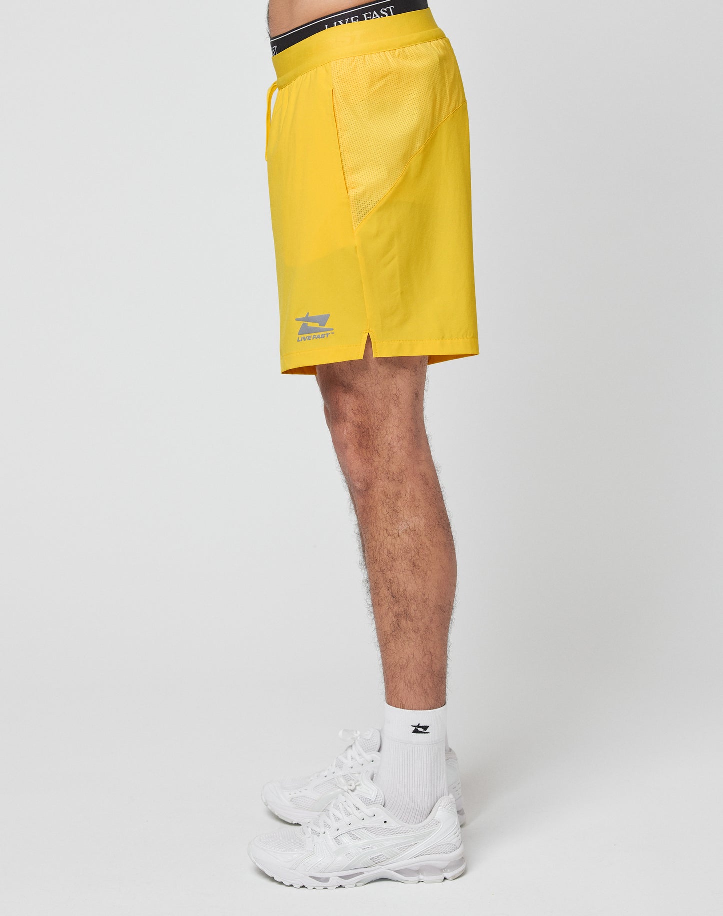 A person models the LIVE FAST DIE YOUNG Live Fast Performance Shorts in bright yellow with an elastic waistband, paired with white socks and sneakers. Only the lower body is visible against a plain white background.