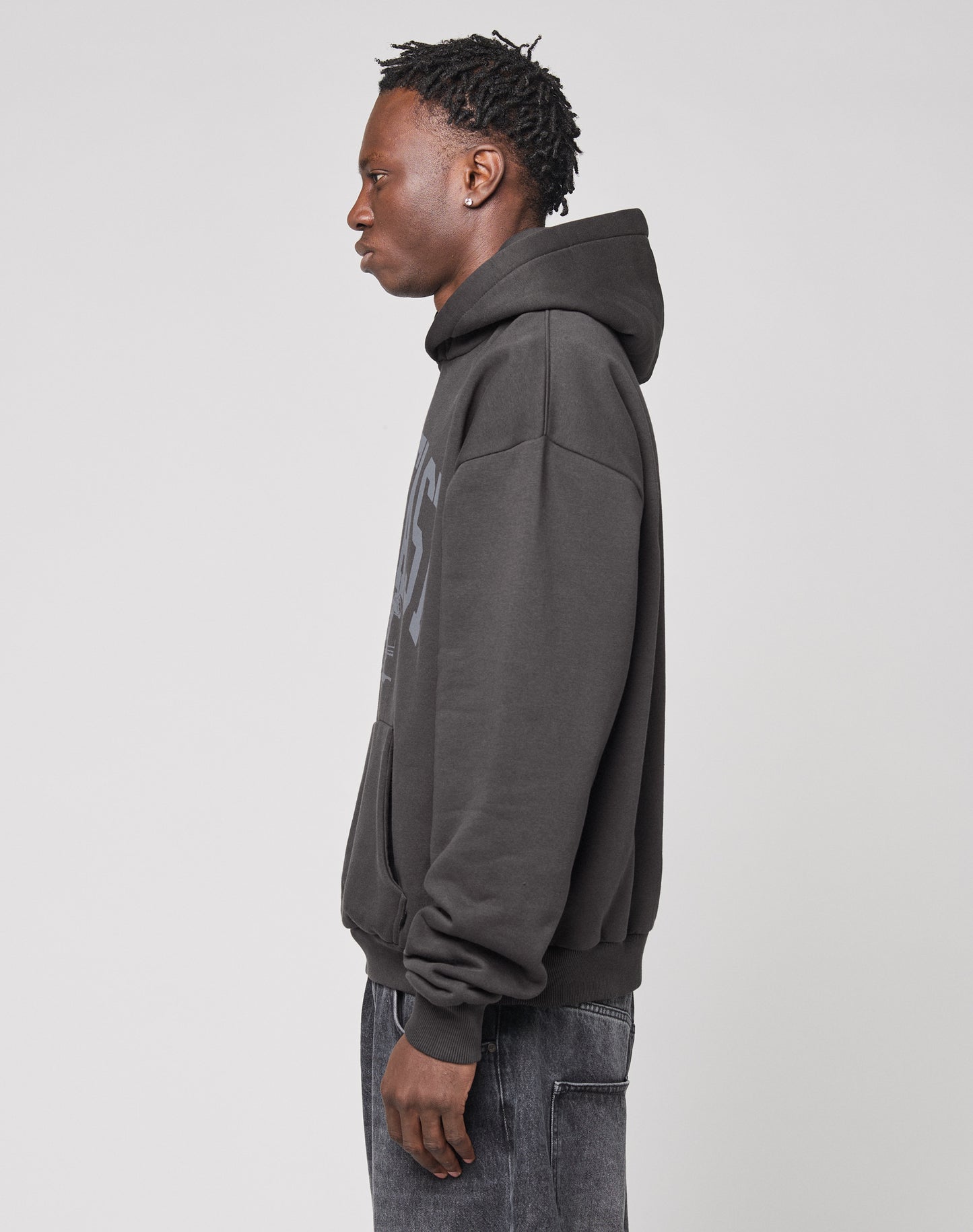 A person with short, textured hair stands in profile against a plain background, wearing the LIVE FAST DIE YOUNG Live Fast Stallion Hooded—a dark, oversized hoodie crafted from soft Baumwolle with an eye-catching front print—and dark jeans.
