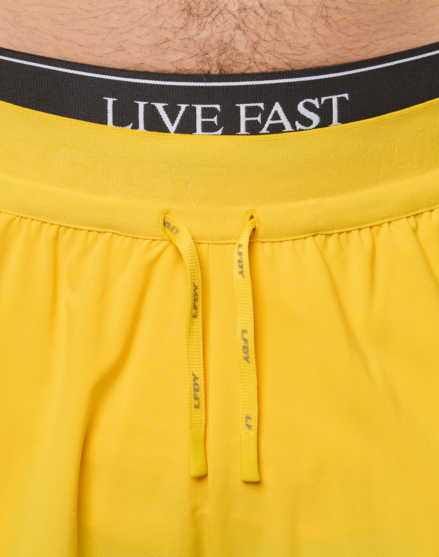 Close-up of a person’s lower torso wearing LIVE FAST DIE YOUNG’s bright yellow Live Fast Performance Shorts with a drawstring, paired with black underwear featuring the words LIVE FAST on the waistband.