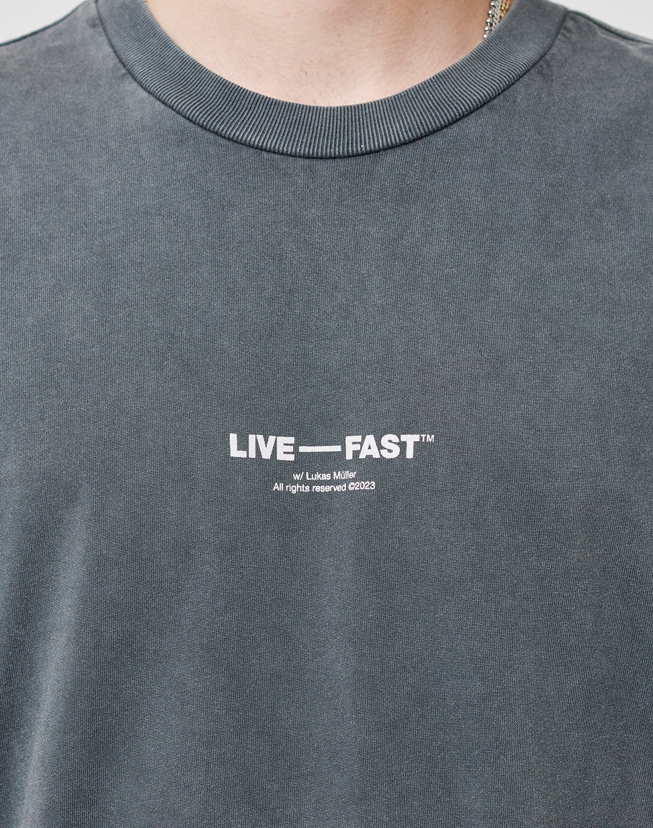 A close-up of someone wearing the Its Vulgar Tee by LIVE FAST DIE YOUNG, an oversized faded dark gray 100% Baumwolle shirt with LIVE FAST™ in white and small text below on the chest. The face is not visible.