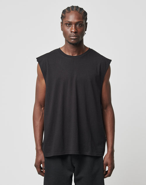 A man with braided hair wears the LIVE FAST DIE YOUNG Basic Muscle Shirt, a sleeveless black top with deep armholes, paired with black pants. He stands against a plain light gray background, looking straight at the camera with a neutral expression.