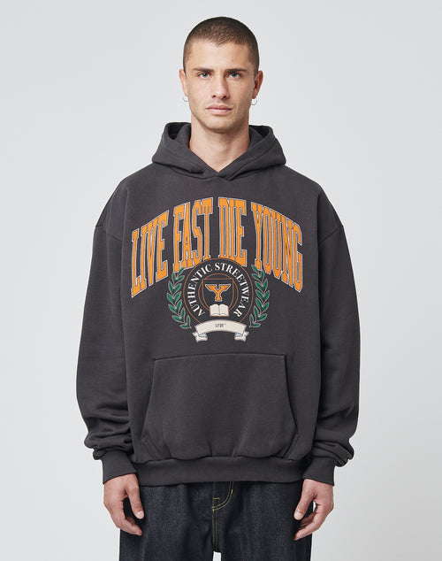 A young man with short hair wears the LIVE FAST DIE YOUNG Live Fast Die Young Hooded, a black hoodie with bold orange “LIVE FAST DIE YOUNG” lettering and laurel graphic, made from soft Baumwolle Polyester, standing before a plain light gray background.