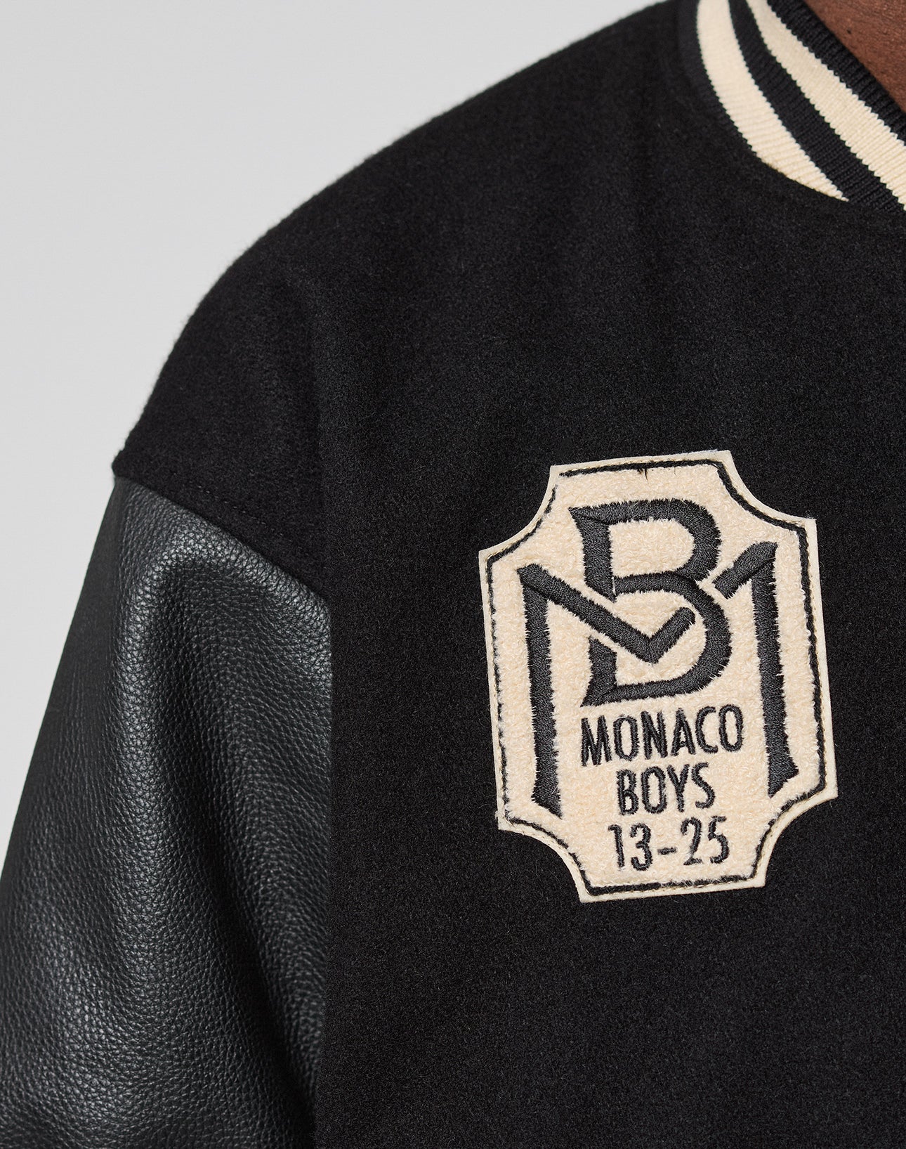 Close-up of the LFDY x Schott NYC Varsity Jacket by LIVE FAST DIE YOUNG, featuring black leather sleeves, a “B1 MONACO BOYS 13-25” chest patch, and classic black-and-white striped collar—signature Schott NYC style.