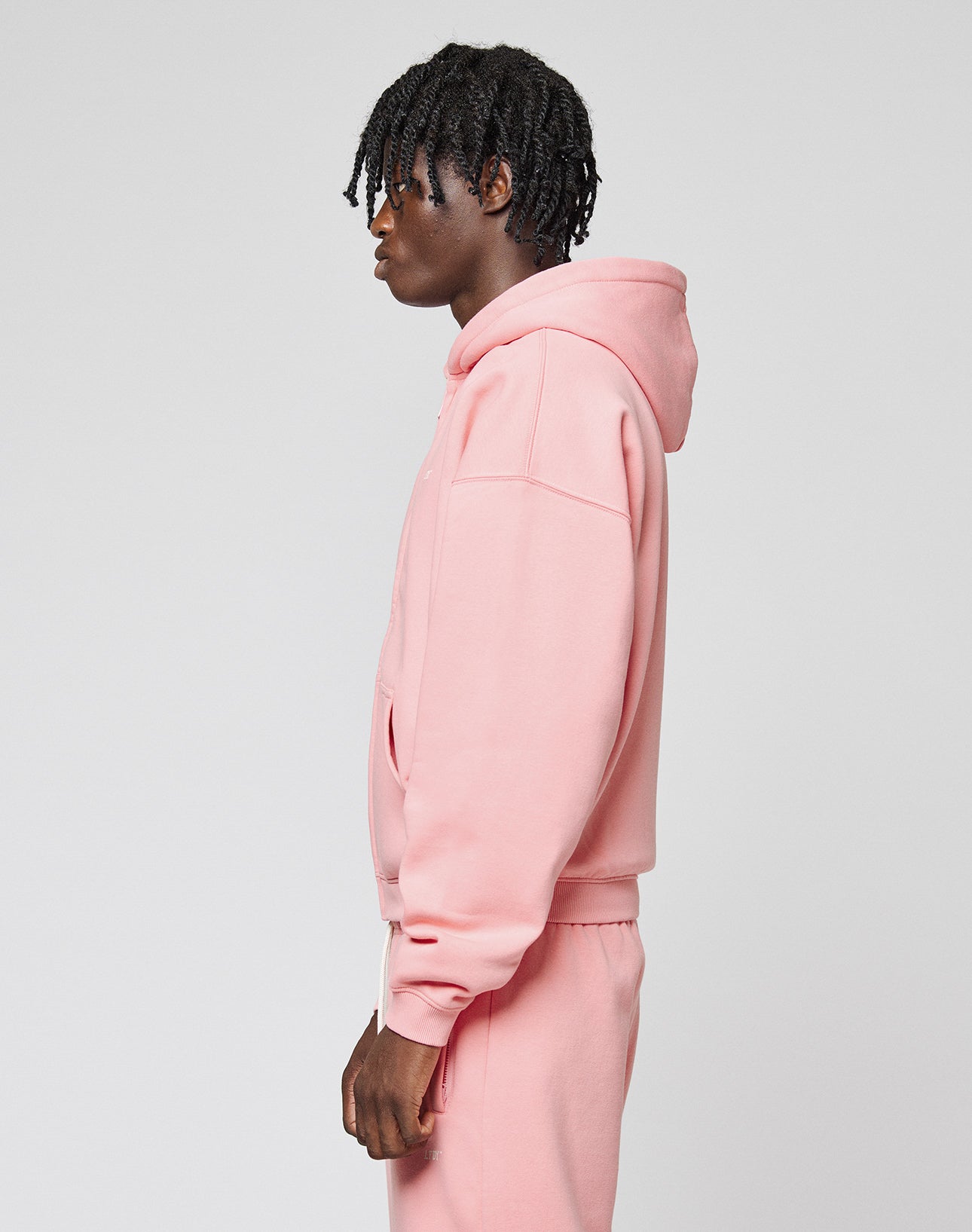 A person with short, curly hair stands in profile, wearing the LIVE FAST DIE YOUNG Basic Hooded Zip in light pink and matching sweatpants against a plain light gray background.
