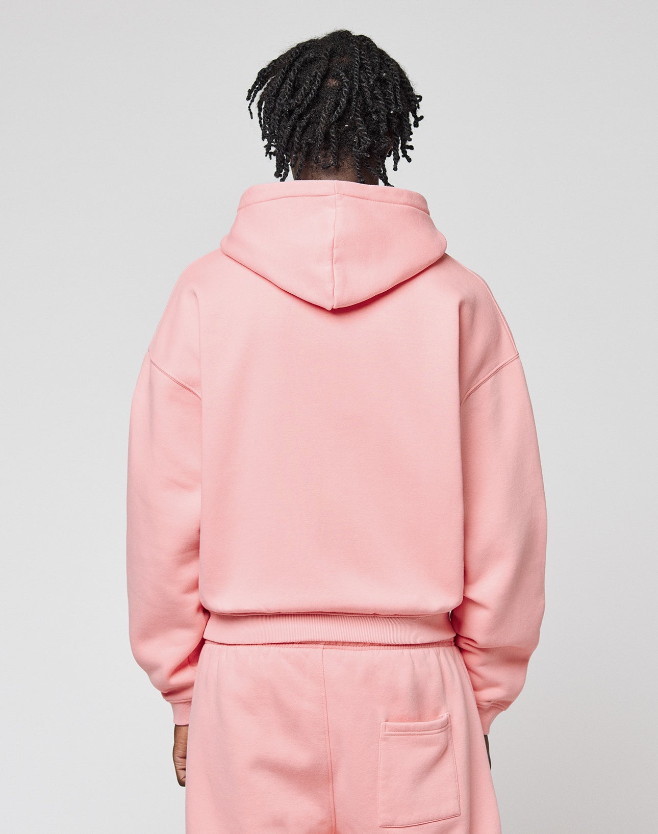 A person with short, twisted hair faces away from the camera, wearing a light pink Basic Hooded Zip and matching sweatpants by LIVE FAST DIE YOUNG, featuring logo branding on the back pocket against a plain light gray background.
