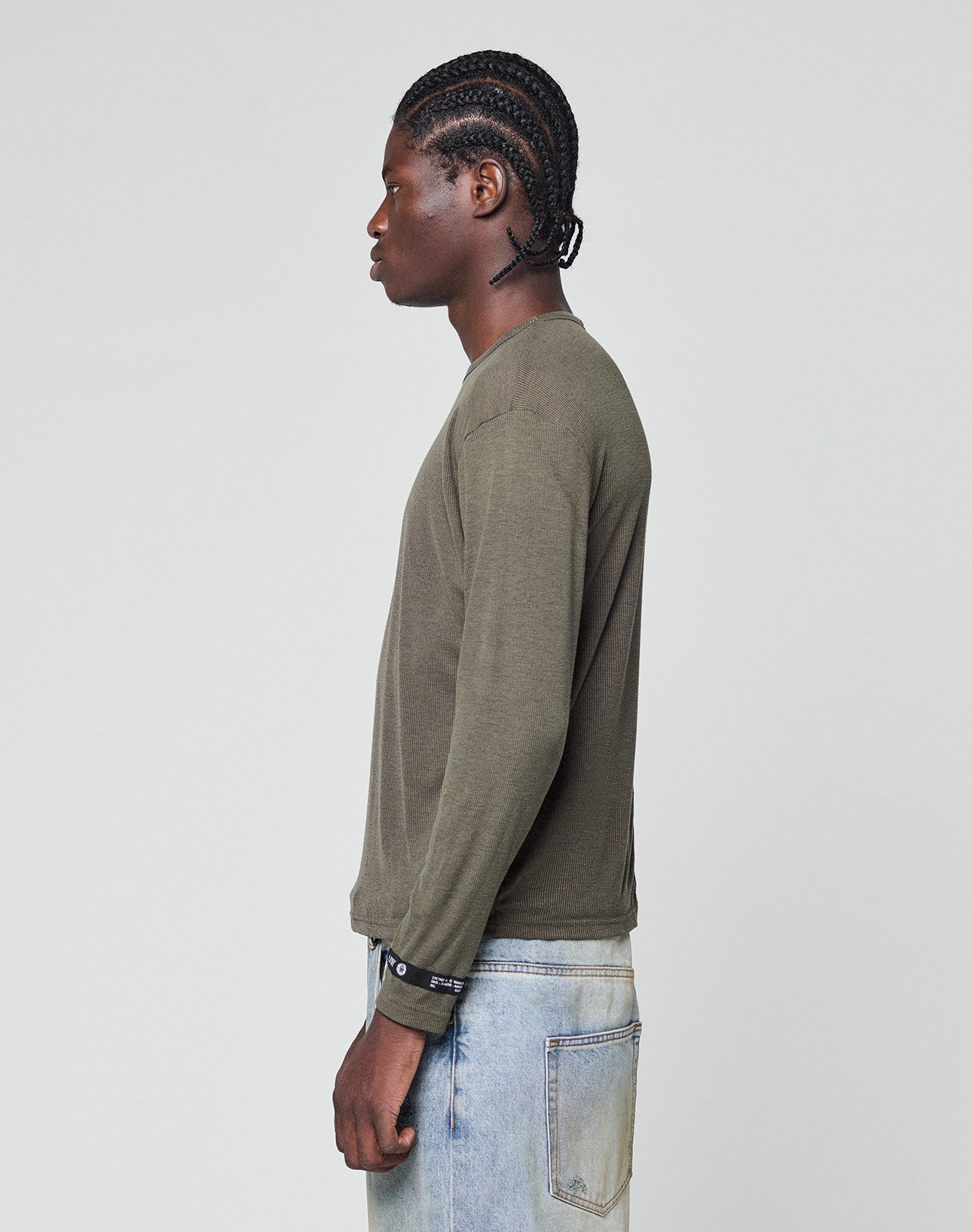 A person with braided hair stands in profile against a plain background, wearing the LIVE FAST DIE YOUNG LFDY Rib Longsleeve in olive green and light blue jeans.