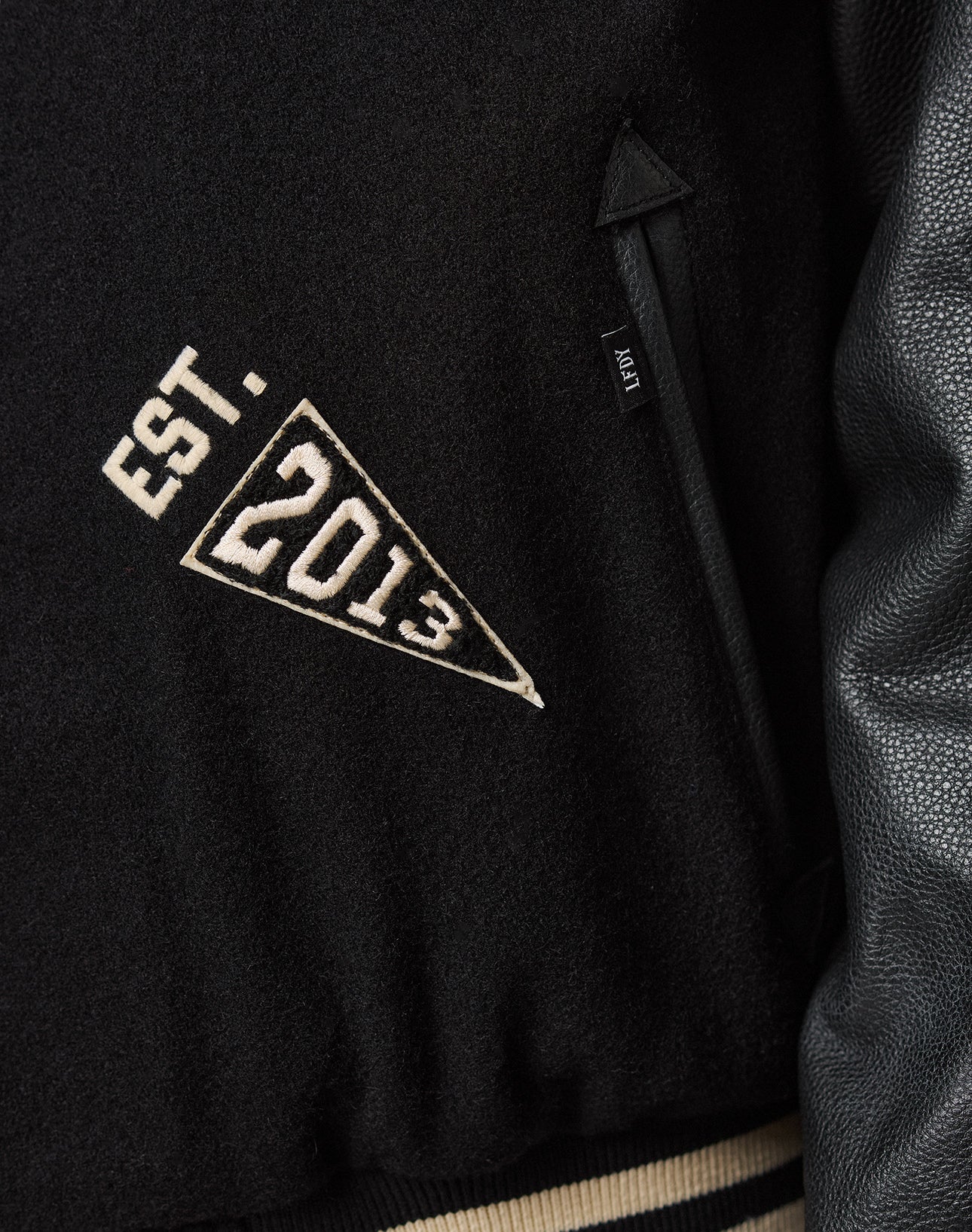 Close-up of the LFDY x Schott NYC Varsity Jacket by LIVE FAST DIE YOUNG, featuring a white EST. 2013 triangular patch and a black FSTY tag near the black leather sleeve, inspired by classic Schott NYC craftsmanship.