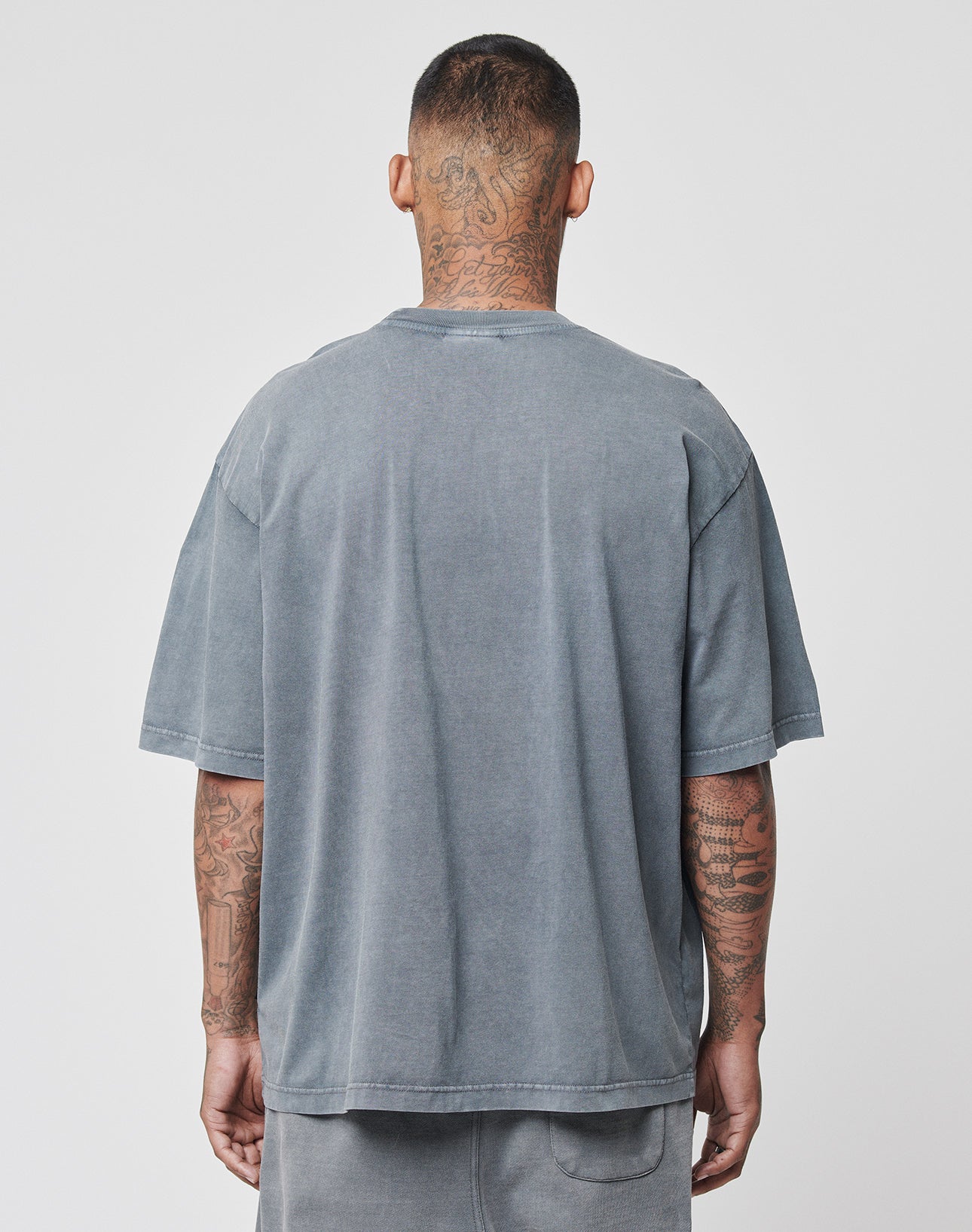A person with short hair and visible tattoos faces away from the camera, wearing the LIVE FAST DIE YOUNG Basic Light Tee in faded gray Oversize Fit and matching vintage-washed shorts, set against a plain background.