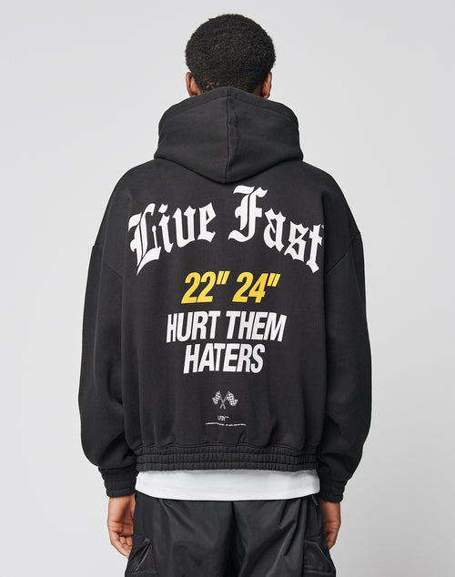 A person wears the LFDY Haters Hooded Zip by LIVE FAST DIE YOUNG, an oversize black hoodie with a double-layer hood and white/yellow Live Fast and 22 24 HURT THEM HATERS text plus small graphics on the back.
