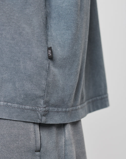 Close-up of a person wearing the LIVE FAST DIE YOUNG Basic Light Tee and matching pants in Oversize Fit. The 100% Baumwolle tee has a small black tag on the side seam. Background is plain and light-colored.