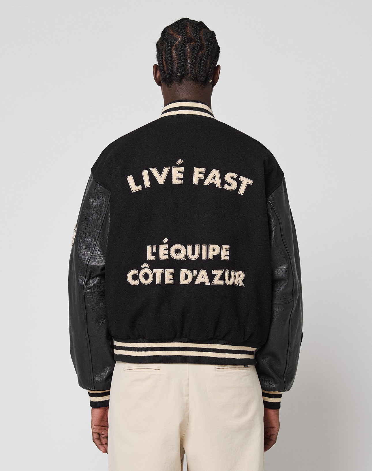 A person with braided hair wears the black LFDY x Schott NYC Varsity Jacket by LIVE FAST DIE YOUNG, featuring leather sleeves and bold beige LIVÉ FAST LÉQUIPE CÔTE D’AZUR text on the back as they face away from the camera.